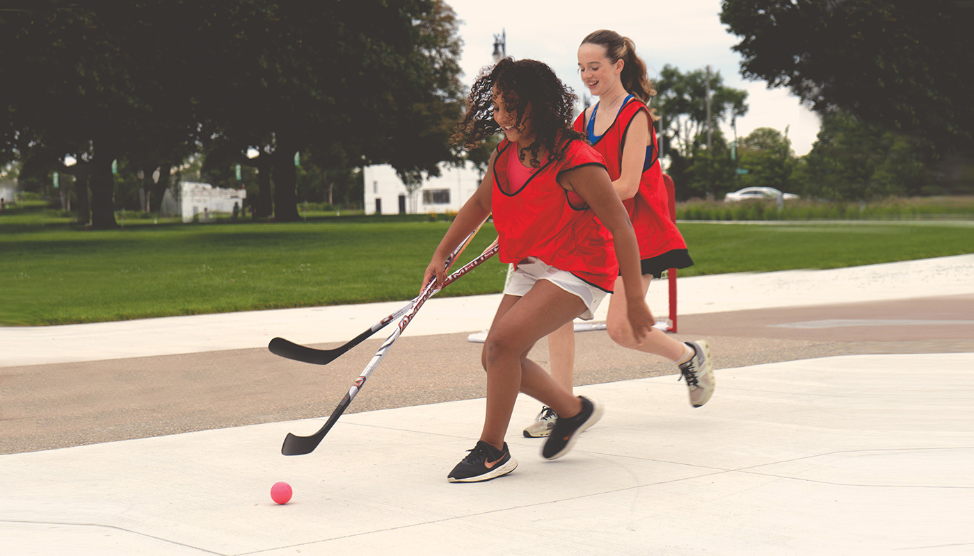 A brown-skinned girl with curly hair and a white girl with a ponytail play street hockey together.