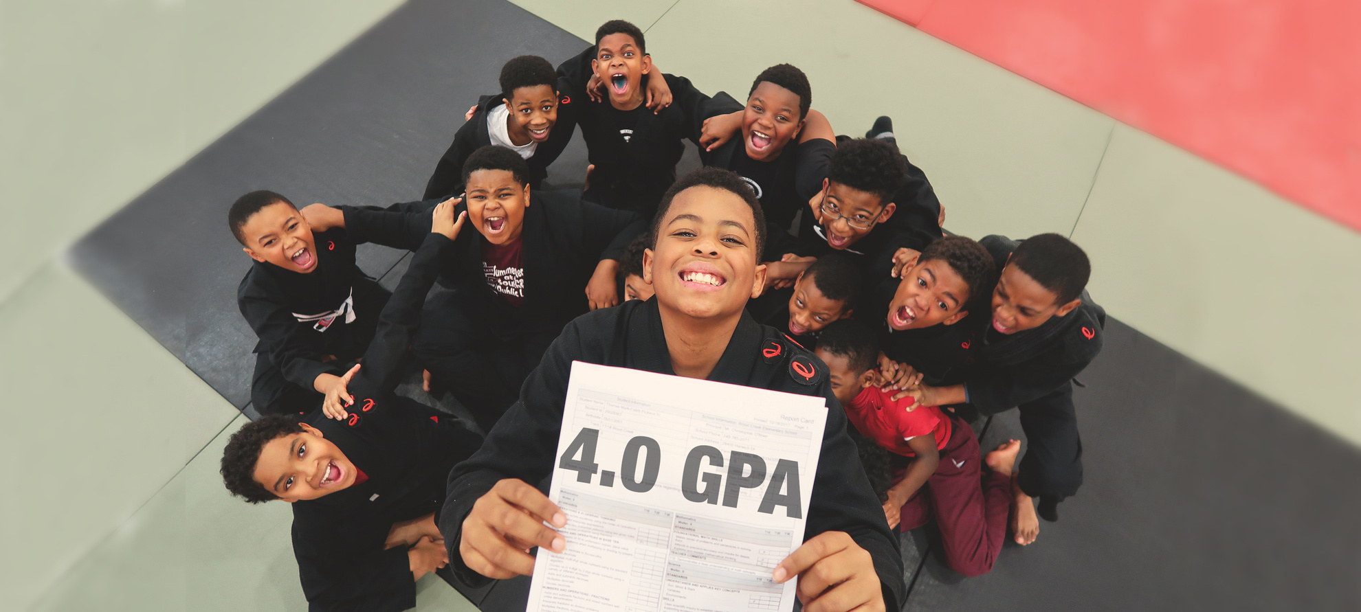 Kids with brown skin, wearing martial arts uniforms, smile and hold up a sign that reads, “four point oh GPA”